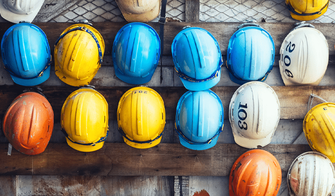 Several rows of brightly coloured hard construction hats hang on a cladded wall.